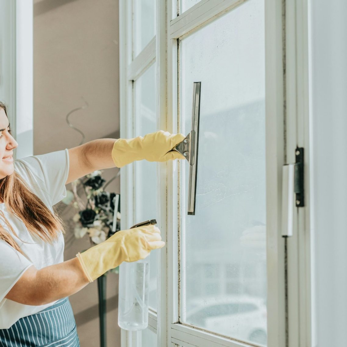 Femme nettoyant une fenêtre avec un racloir, portant des gants en caoutchouc jaunes.